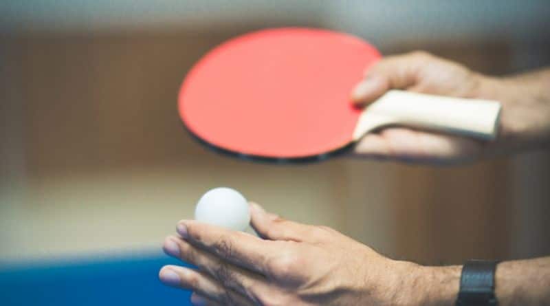 Tischtennisschläger mit rotem Belag wird in einer Hand gehalten, in der anderen ein Tischtennisball
